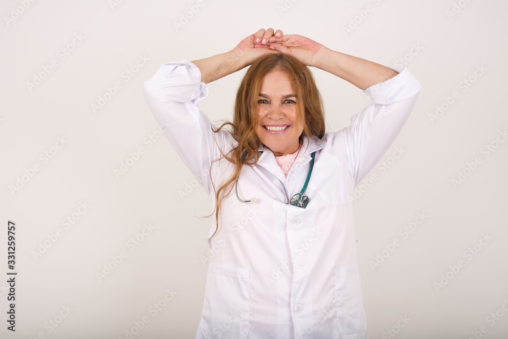 Portrait of beautiful brunette doctor female looks with excitement at camera, keeps hands raised over head notices something unexpected, isolated over gray wall. Lovely woman reacts on sudden news. Portrait of beautiful brunette doctor female looks with excitement at camera, keeps hands raised over head notices something unexpected, isolated over gray wall. Lovely woman reacts on sudden news.