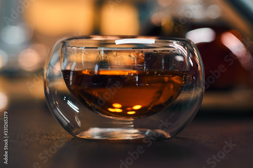 Transparent bowl with black tea on a wooden table. Blurred background of bright lights. Horizontal picture.