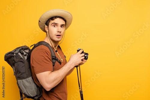 shocked traveler in hat with backpack holding photo camera on yellow
