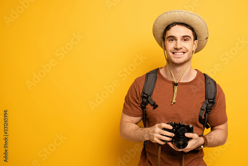 cheerful traveler in hat with backpack holding photo camera on yellow © LIGHTFIELD STUDIOS