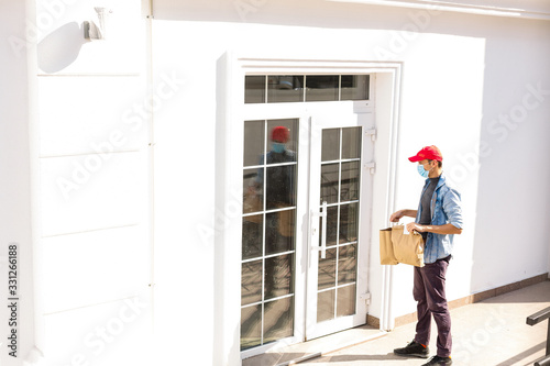 Delivery man holding paper bag with food on white entrance of house background , food delivery man in protective mask © Angelov
