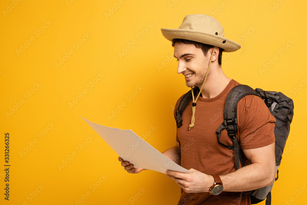 handsome smiling tourist in hat with backpack looking at map on yellow handsome smiling tourist in hat with backpack looking at map on yellow