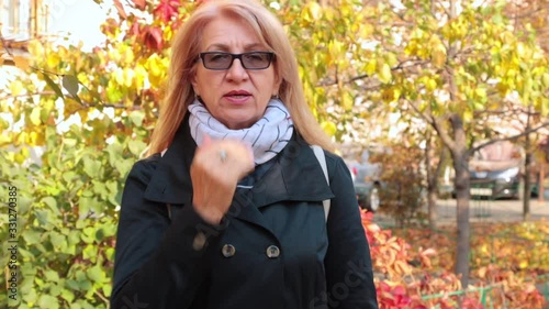 Adult long-haired blonde woman in trench coat sends a kiss. Outdoors. Older people and communication conceptt.