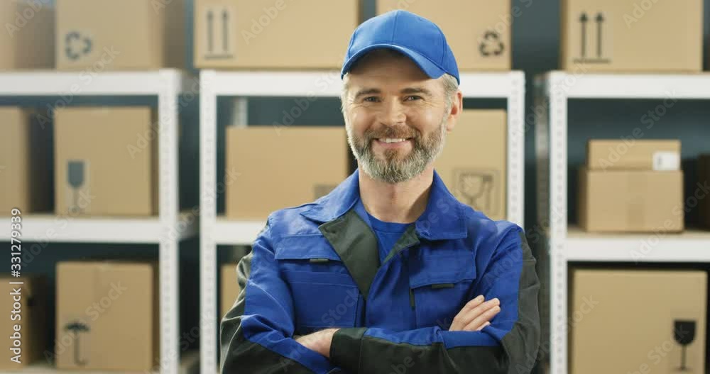Portrait of Caucasian handsome deliveryman with gray beard in blue uniform and cap smiling cheerfully to camera in postal office store among shelves with parcels. Man mail courier standing in storage. Portrait of Caucasian handsome deliveryman with gray beard in blue uniform and cap smiling cheerfully to camera in postal office store among shelves with parcels. Man mail courier standing in storage.