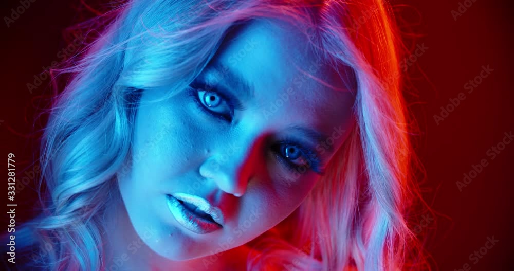 Sexy caucasian blonde girl look at camera with sensual stare, weariing bright glowing makeup, standing in red and blue neon lights - nightlife, cybcerpunk concept 4k footage close up portrait Sexy caucasian blonde girl look at camera with sensual stare, weariing bright glowing makeup, standing in red and blue neon lights - nightlife, cybcerpunk concept 4k footage close up portrait
