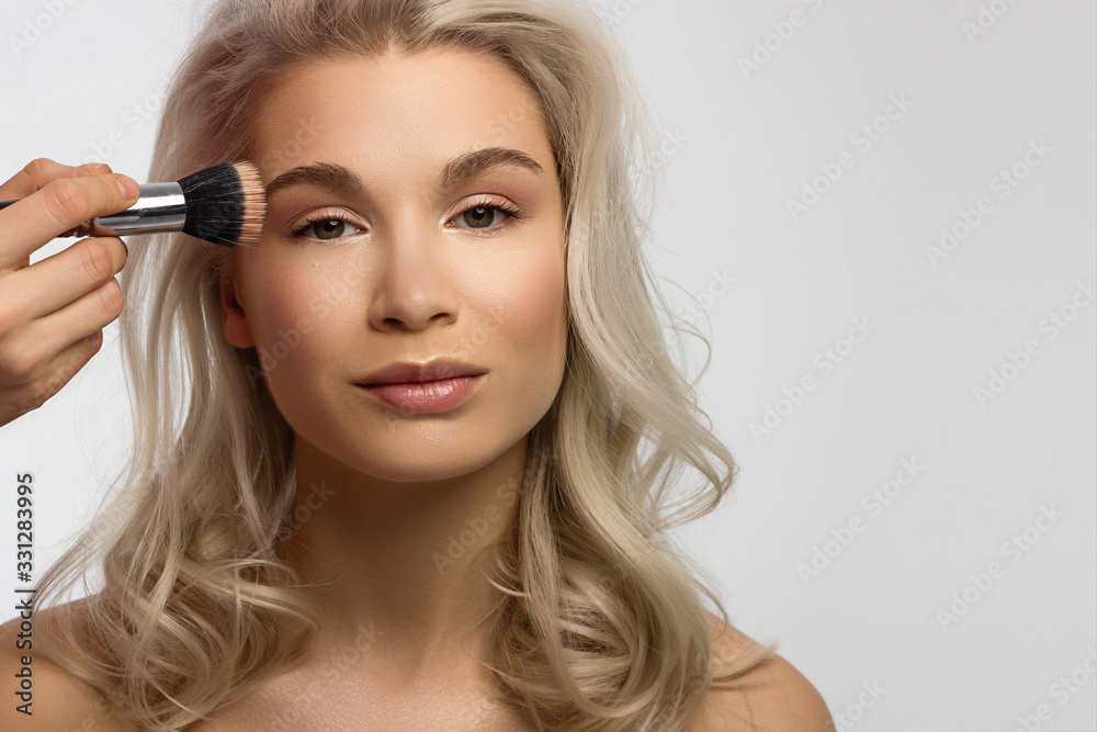 Beauty close-up of a blond woman applying powder to clean her face skin. Looks at the camera with a sexy look. Soft skin care, natural makeup. Pink lip gloss, smooth and thick eyebrows. curly hair