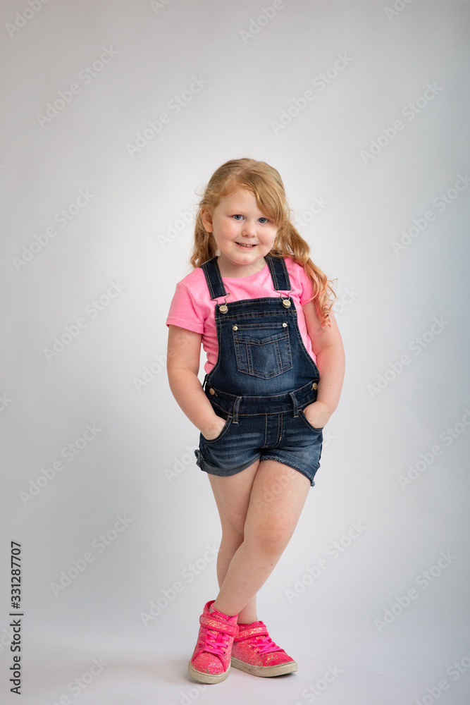 Full body portrait of young blond female girl crossed legs hands in jean overall pockets sweet smile 