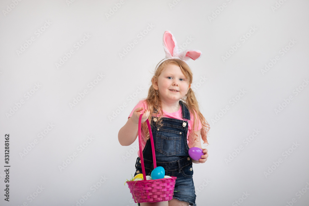 Small little girl holding up a pink Easter egg basket wearing white bunny ears excited happy expression 