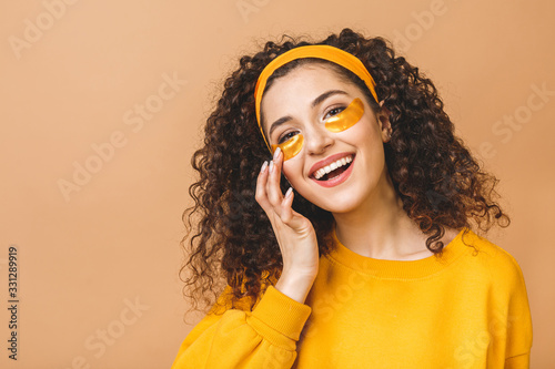 Photo Image of young pure beautiful curly woman isolated over beige background take care of her skin with under eye patches