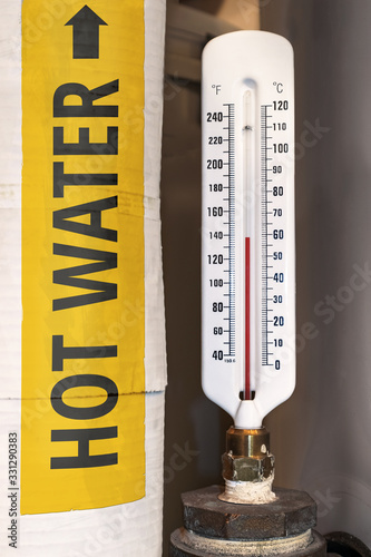 Thermometer close up of hot water tank in boiler room of a residential or strata building. Yellow label "hot water" with arrow on insulated white pipe.