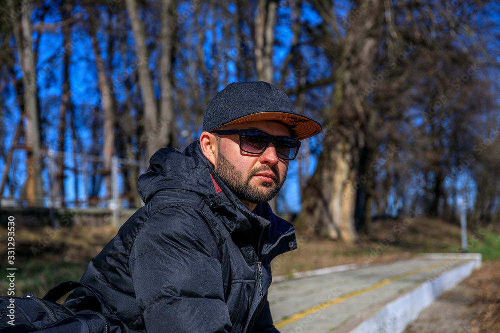 Stylish young man with a cap, sunglasses, sneakers in a denim jacket with a backpack sits on golden foliage in the park. Attractive hipster guy is rests at the forest. Stylish young man with a cap, sunglasses, sneakers in a denim jacket with a backpack sits on golden foliage in the park. Attractive hipster guy is rests at the forest.