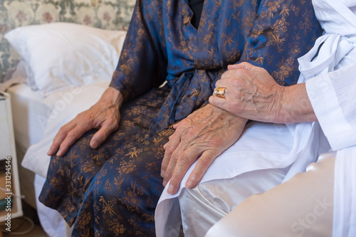 Close up cropped view of elderly man and woman's hands while sitting on bed in robes - coronavirus isolation concept (selective focus)