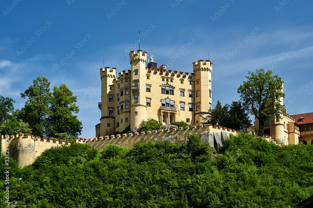 A view of a Hoehenschwangau castle on top of a hille on a sunny summer day against a blue sky and green foliage bellow the castle wall.
