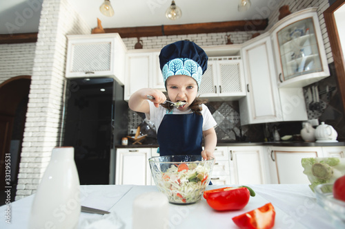 Child in an apron and a chef's hat is eating a vegetable salad in the kitchen and looking at the camera.