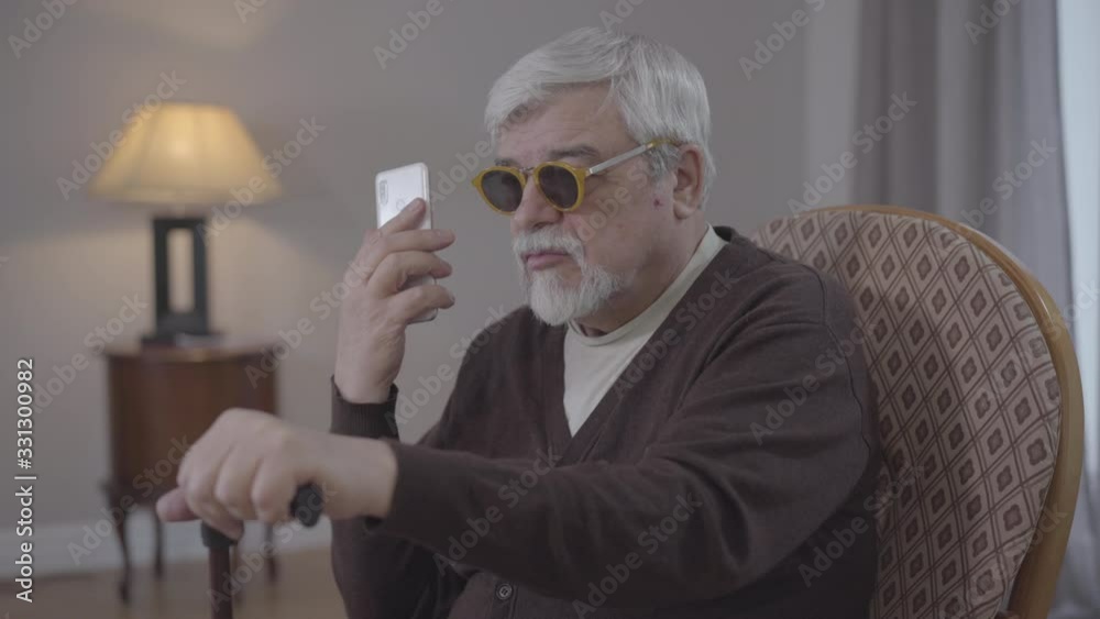 Portrait of blind old Caucasian man talking on the phone indoors. Grey-haired male retiree in armchair holding smartphone and chatting. Modern technologies for disabled people, lifestyle. Portrait of blind old Caucasian man talking on the phone indoors. Grey-haired male retiree in armchair holding smartphone and chatting. Modern technologies for disabled people, lifestyle.