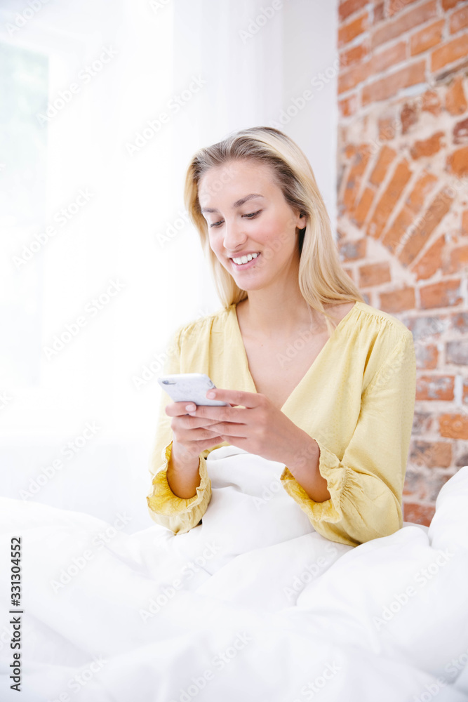 Beautiful blonde woman wearing pajama using phone in her bedroom. Beautiful blonde woman wearing pajama using phone in her bedroom.