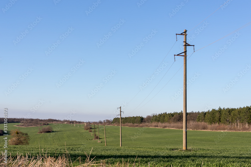 A line of electric poles with cables of electricity in a field with a forest in background in spring time during the day.