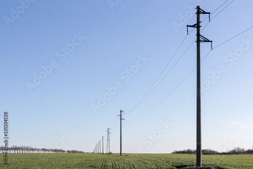 A line of electric poles with cables of electricity in a field with a forest in background in spring time during the day.