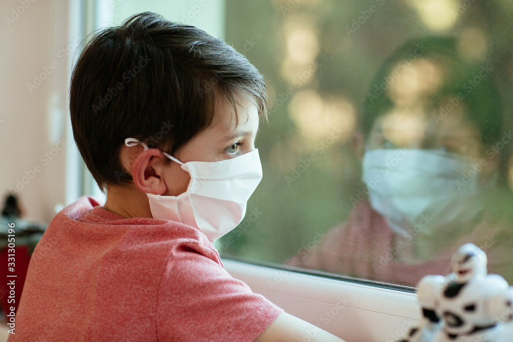 Children and coronavirus. Close up of child with protective face mask looking outside window.