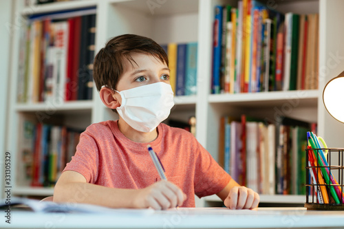 Young student wearing protective mask working in isolation on school assignments. Child wearing face mask self-studying at home during coronavirus outbreak.