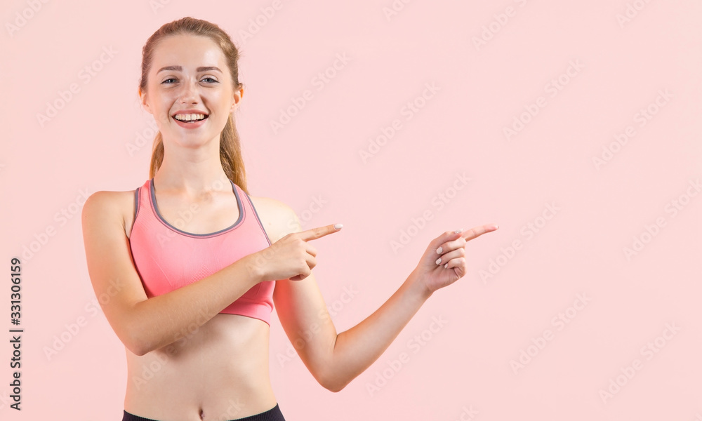Young athletic woman points finger to the side right while standing on a light pink background. Cheerful sportswoman pointing on copy space. Young athletic woman points finger to the side right while standing on a light pink background. Cheerful sportswoman pointing on copy space.