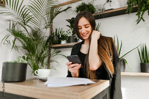 Beautiful young businesswoman sitting in a cozy cafe with a cup of coffee and documents, uses a smartphone with a smile on her face. Attractive happy girl in a business suit uses the internet © bodnarphoto