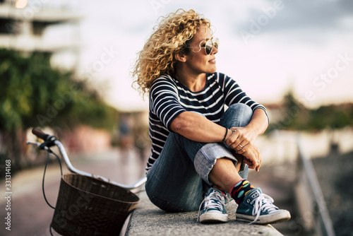 Beautiful adult caucasian young woman with blonde curly hair enjoy the outdoor leisure activity relaxing and sitting on a wall with bike in background - people portrait with sky in background