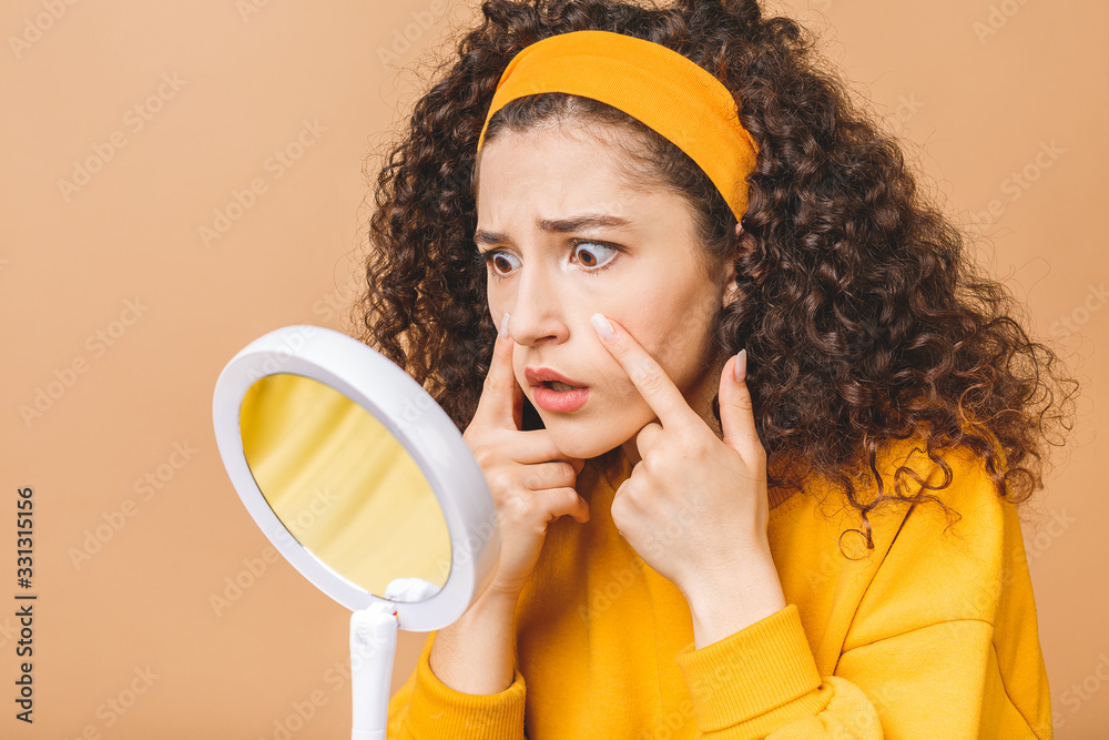 Φωτογραφία Beauty portrait of an astonished shocked amazed attractive curly woman examining her face while holding a mirror isolated over beige background