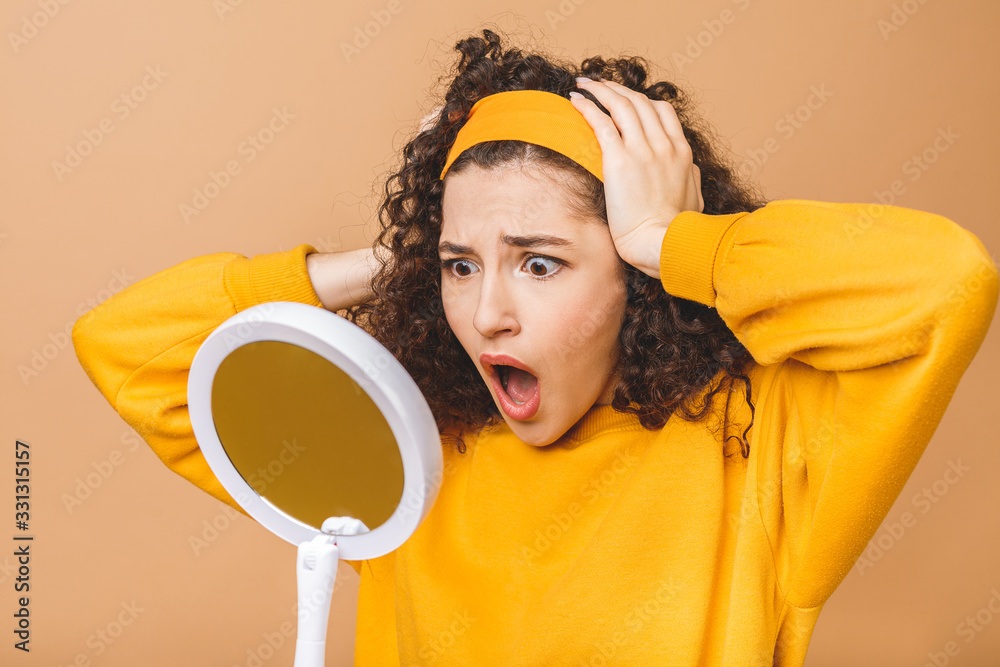 Φωτογραφία Beauty portrait of an astonished shocked amazed attractive curly woman examining her face while holding a mirror isolated over beige background