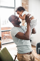 Joyful Afro American man lifting cute baby up in the air