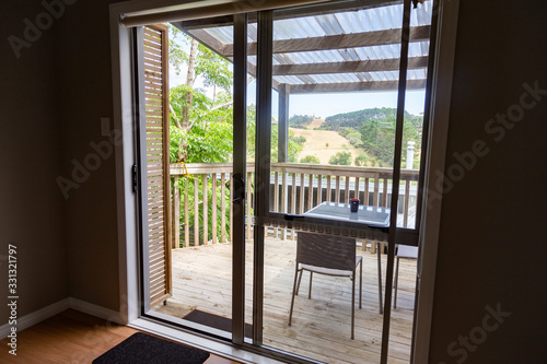 Looking out to the deck of a small house in rural west Auckland. View of rolling hills, dry grass in summer. Small table and chairs. Through sliding glass door. Peaceful and quiet countryside cottage