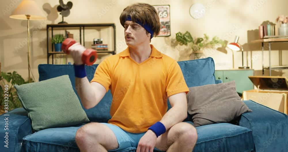 Smiling Caucasian athlete training biceps while sitting on blue couch in living room. Retro style sportsman doing exercise with red dumbbell at home. Joyful male practicing sport indoors. 60s concept Smiling Caucasian athlete training biceps while sitting on blue couch in living room. Retro style sportsman doing exercise with red dumbbell at home. Joyful male practicing sport indoors. 60s concept