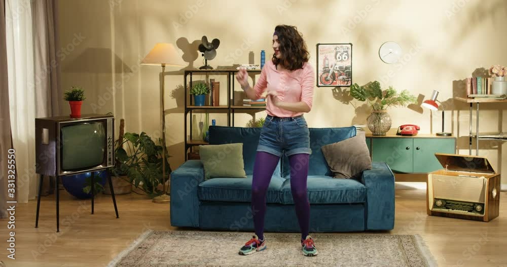 Young pretty funny Caucasian retro style girl dancing at blue sofa in living room at home. Joyful smiling athlete female model fooling around while posing to camera. 50s concept Young pretty funny Caucasian retro style girl dancing at blue sofa in living room at home. Joyful smiling athlete female model fooling around while posing to camera. 50s concept