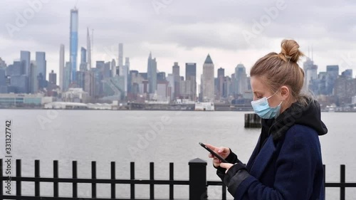 Coronavirus pandemic. Woman is wearing protection mask using smart phone on New York background