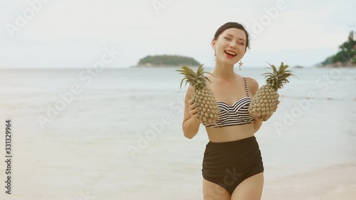 Beach fun. Happy young woman wearing swimsuit laughing and smiling with two pinapples. Cheerful asian girl with tropical fruit at sea. Summertime, vacation, lifestyle, nature, natural concept. photo