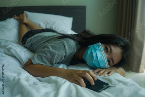 pandemic and virus outbreak - young beautiful scared and worried Asian Chinese woman in medical mask checking online news with mobile phone on bed lockdown at home