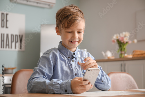 Cute little boy with mobile phone drinking water in kitchen