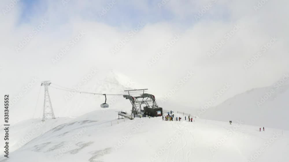 Cable Car/ Lift arrives at a Station on Top of a Mountains. People are standing in front. Diffuse sunlight shines through the clouds. Aerial shot Cable Car/ Lift arrives at a Station on Top of a Mountains. People are standing in front. Diffuse sunlight shines through the clouds. Aerial shot