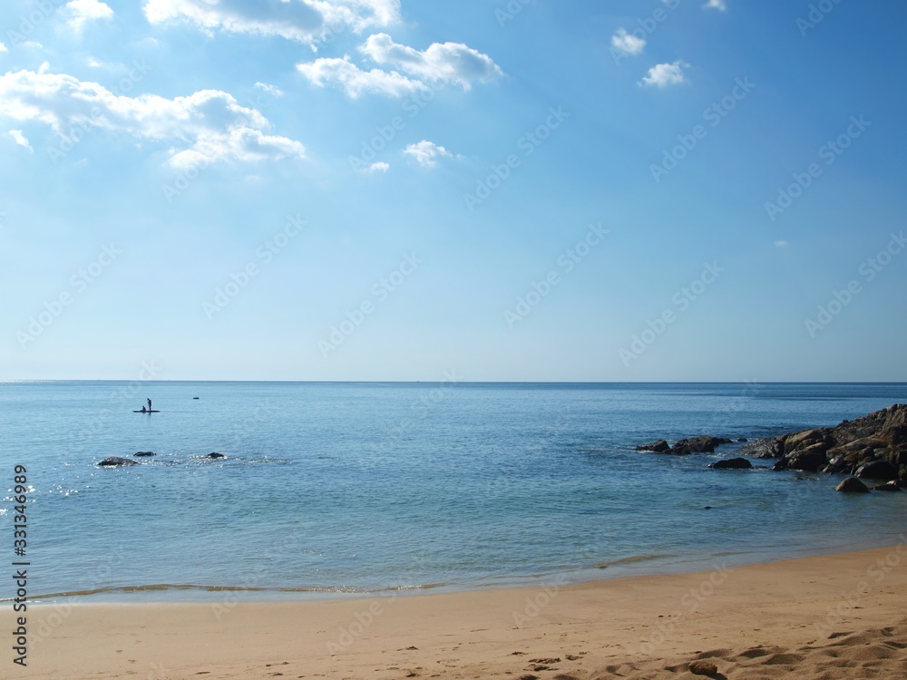 Beach, sea, sand, stones, sun, water surface, horizon, a man is sailing in a boat on oars in the distance. Tropical island paradise on a quiet clear day.