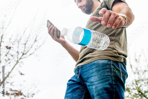 A tan brown haired casual man walking through the park playing on his phone has no regard for the earth and its habitats as he drops his empty plastic water bottle to the ground paying no attention.