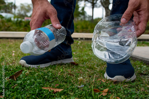 Close up of a man's hands picking up a plastic water bottle and a clear container thrown on the grass to help preserve the world we live from pollution on a nice overcast day.