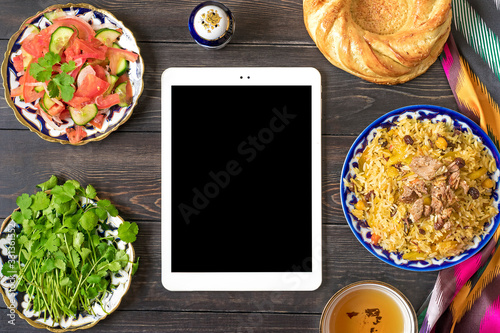 National Uzbek pilaf with meat in plate with traditional pattern, ikat fabric, tablet on wooden table Top view Online food order, delivery concept Flat lay Top view Mockup