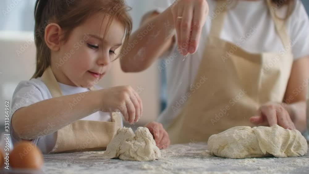 Mother teaching daughter to knead dough for cookies on kitchen in slow motion. Mom teaching kid daughter learning kneading dough with rolling pin, funny child girl helping mother preparing cookies.