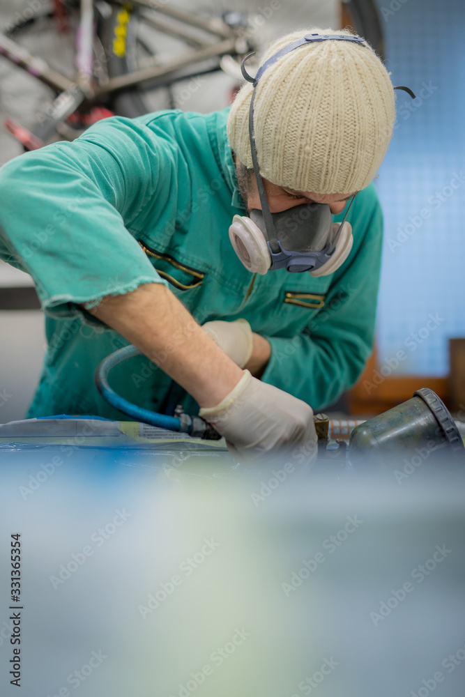 Man in green overalls preparing an engine bay of a car for a final coat of paint. Man spraying grey liquid putty or stucco on the car panels as a last layer before final paint coat. Man in green overalls preparing an engine bay of a car for a final coat of paint. Man spraying grey liquid putty or stucco on the car panels as a last layer before final paint coat.