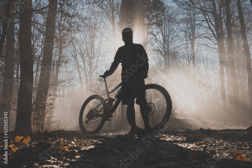 MTB or mountain biker posing in misty forest with sun behind him. Silhouette of a mountain biker in the foggy woods, beautiful sport silhouette