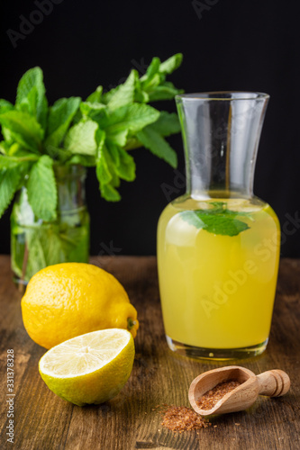 View of lemon jug with slices and mint on dark wooden table with lemon and brown sugar in vertical, on black background