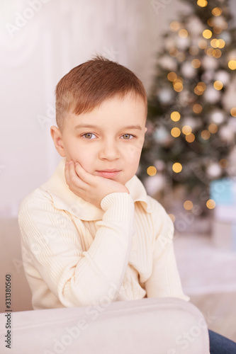 Christmas happy boy. Luxury attractive child. Xmas miracle. Green tree, lights, bokeh, new year interior. Hand near face. Look at camera. Vetrical portrait photo