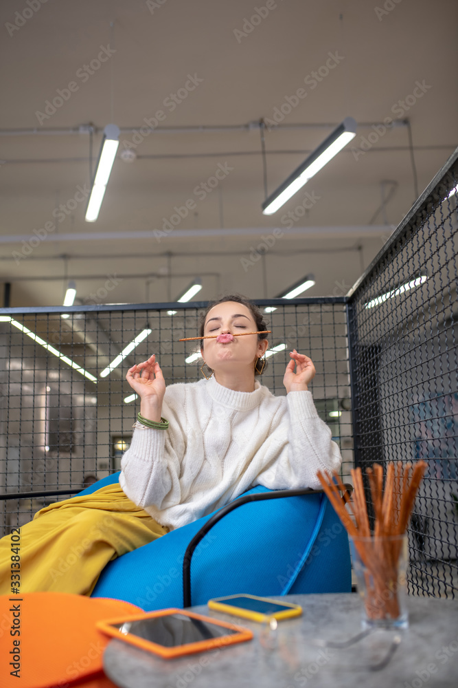 Young woman sitting on bag chair, holding bread stick between lips and nose