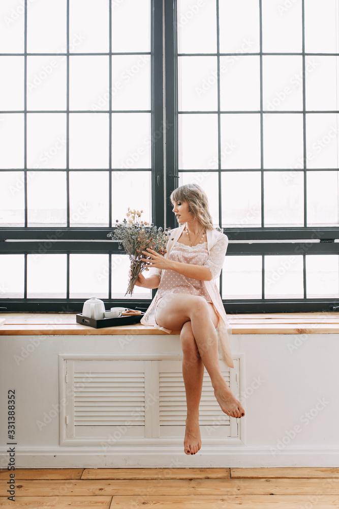 A romantic beautiful attractive sensual sweet girl in a housecoat lies resting on the windowsill , rejoices and sniffs spring flowers against the background of a huge window in a modern house A romantic beautiful attractive sensual sweet girl in a housecoat lies resting on the windowsill , rejoices and sniffs spring flowers against the background of a huge window in a modern house