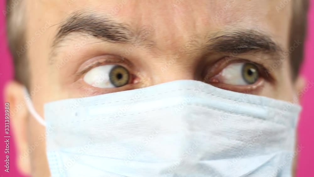 close-up front view on male face in medical mask, looks around with fear and distrust then close eyes in panic, quarantine or epidemic of virus or protection of pollution, pink background close-up front view on male face in medical mask, looks around with fear and distrust then close eyes in panic, quarantine or epidemic of virus or protection of pollution, pink background